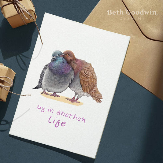 Card with two kissing birds illustration and text 'us in another life' on a blue surface with brown envelopes.