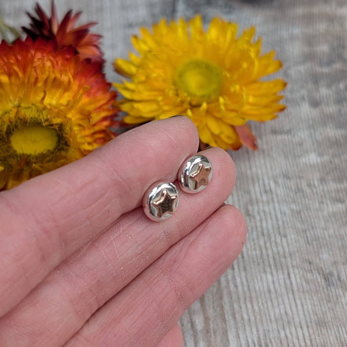A pair of small silver stud earrings with star-shaped cutouts, held on a person's fingers with yellow flowers and a gray wood surface in the background.