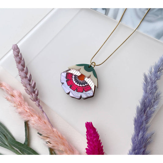 A gold necklace with a flower-shaped pendant featuring a colorful, fan-like pattern in pink, red, green, and white, styled on a white surface with dried pink and purple grasses.