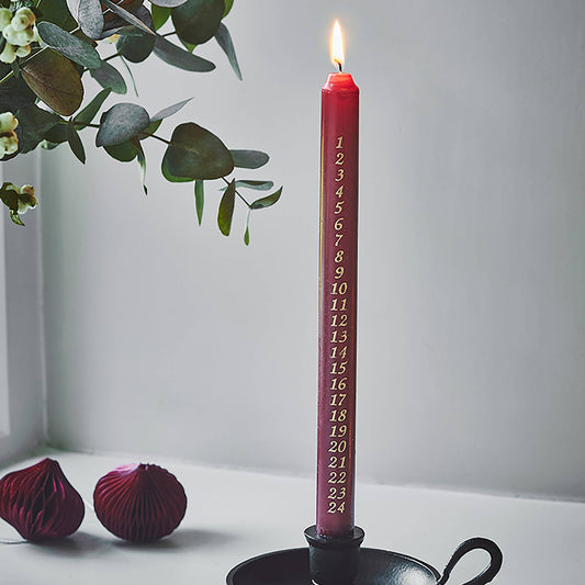 A tall red taper candle in a black holder, lit and standing on a white surface. The candle is printed with the number 24 in gold down the center.
