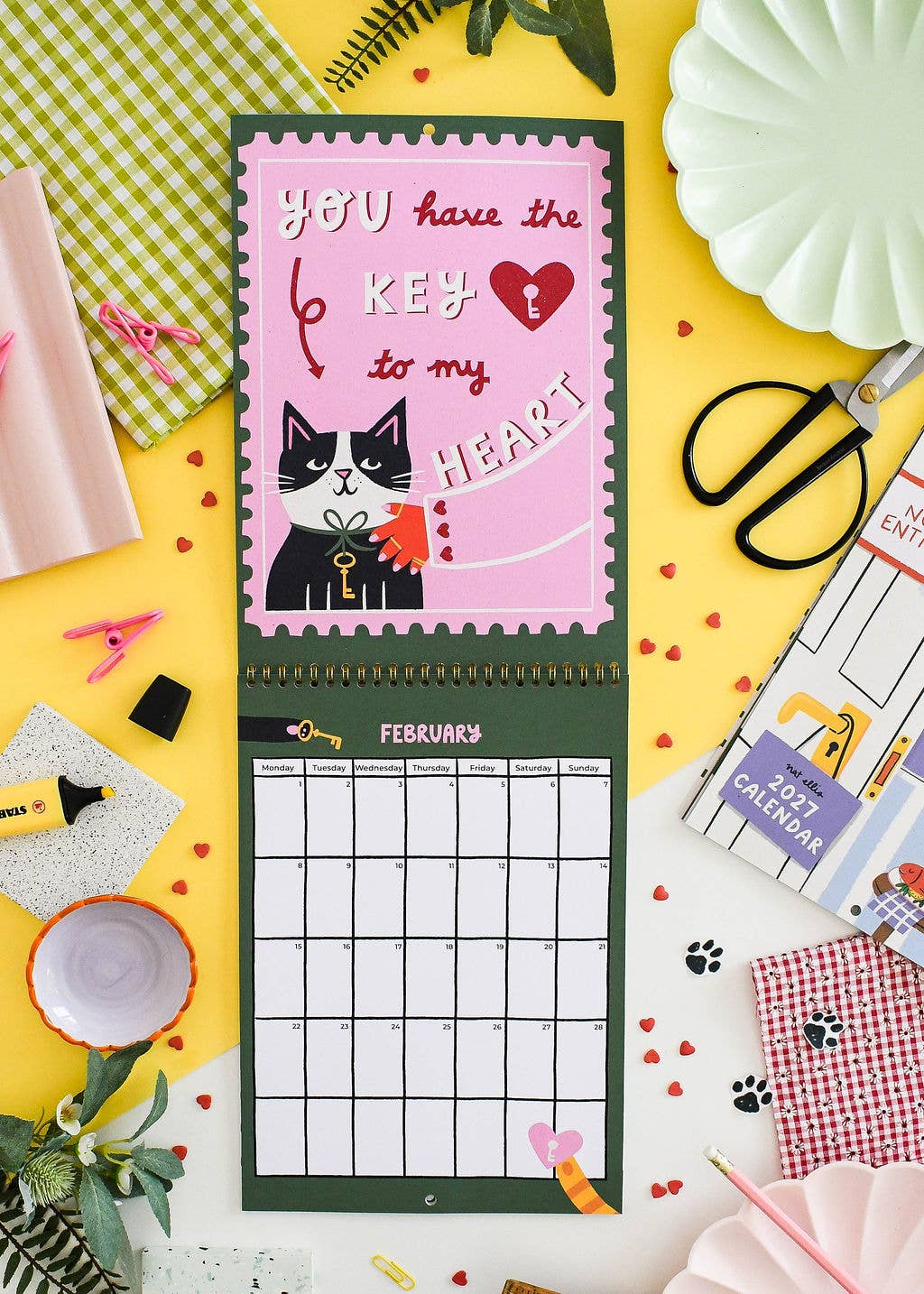 A vertical wall calendar with a pink cat-themed cover reading 'you have the KEY to my HEART' and a February grid below, styled on a yellow surface with craft props, paper fans, scissors, and small heart decorations.