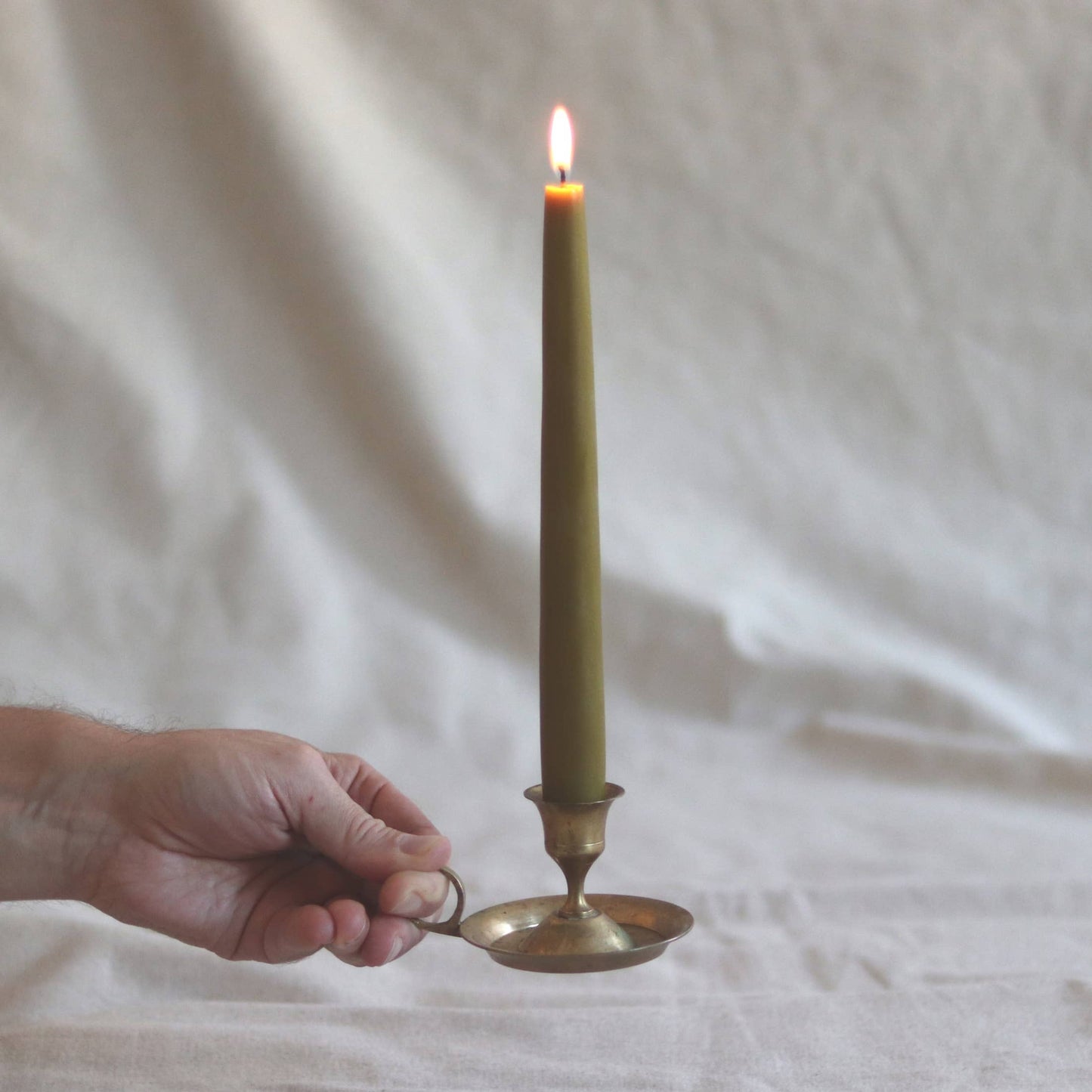 Hand holding a brass candle holder with a lit green candle against a white fabric background