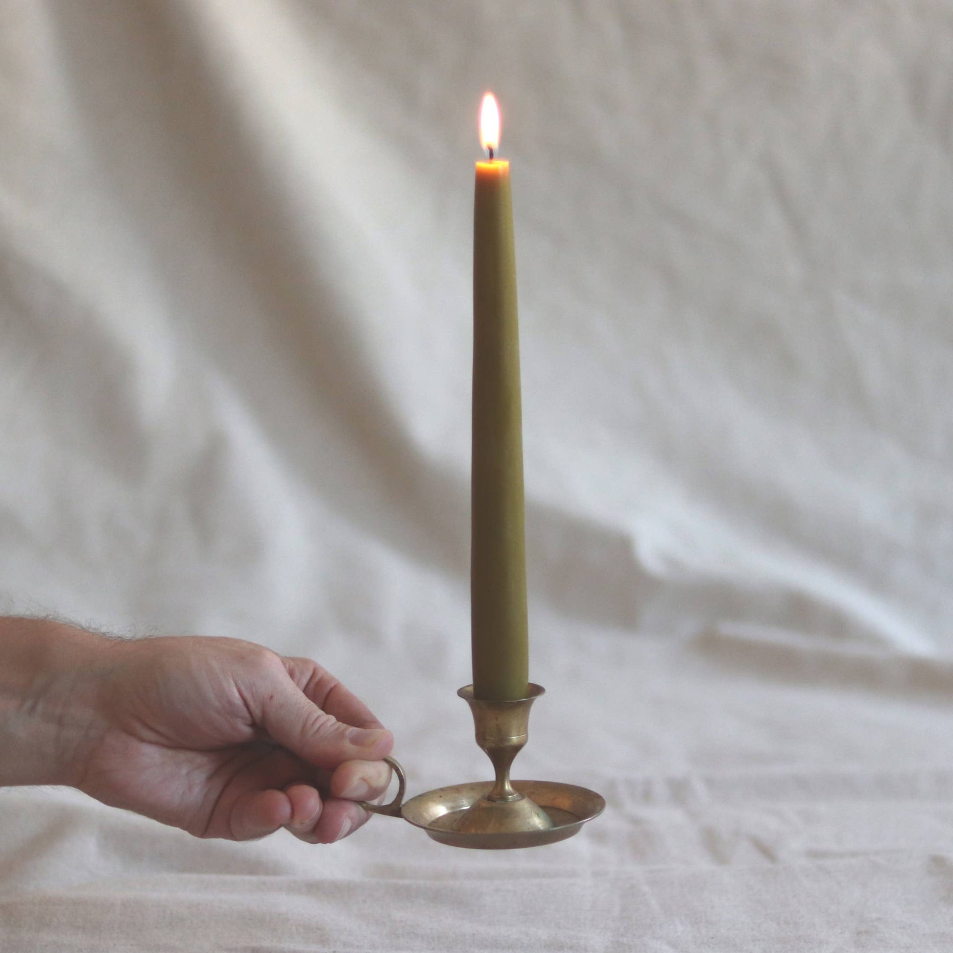 Hand holding a brass candle holder with a lit green candle against a white fabric background