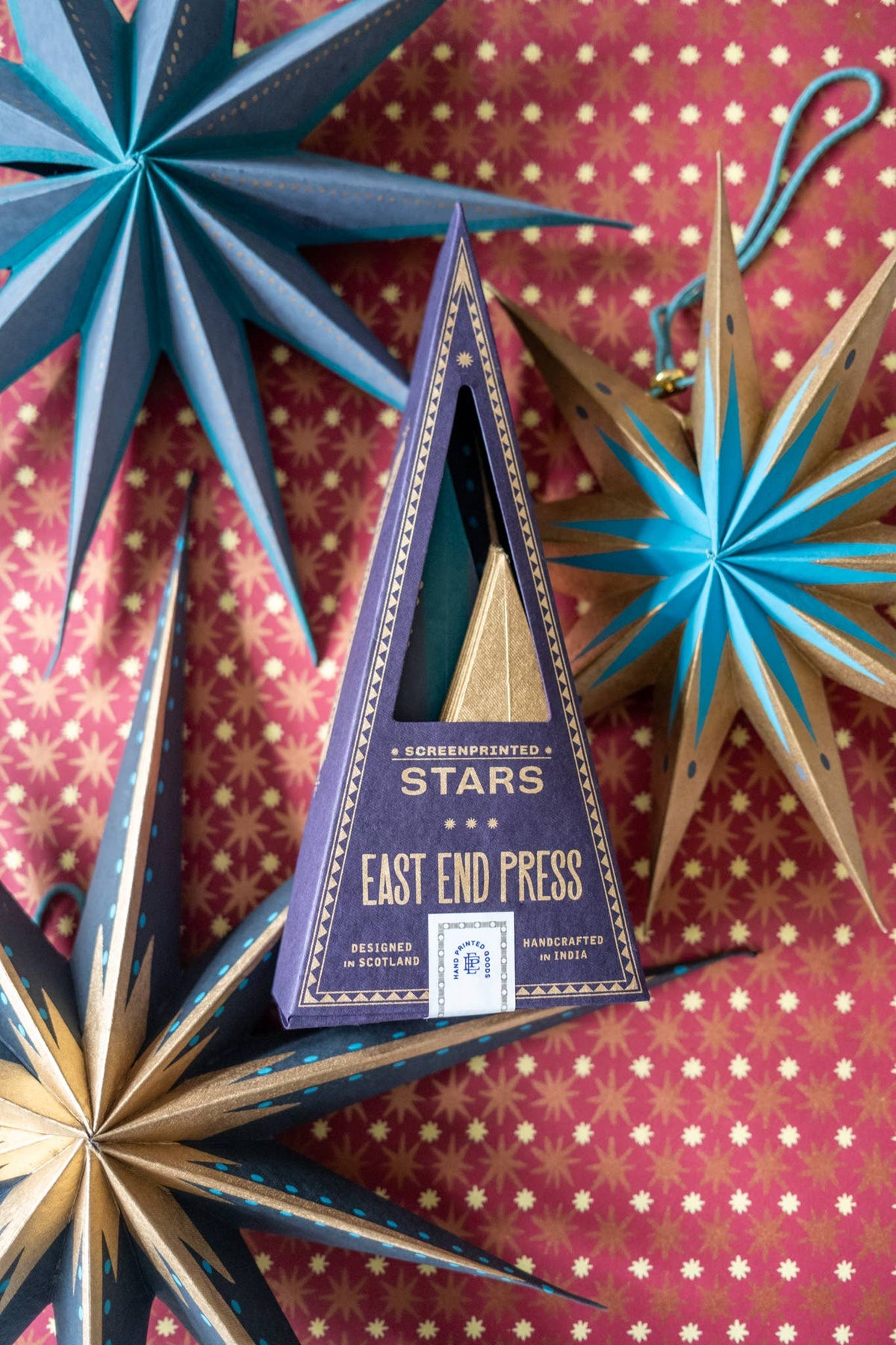 A triangular box labeled 'EAST END PRESS' containing a small gold cone-shaped item, styled among decorative blue and gold star ornaments on a pink patterned background. The box text includes 'CONFEPTUALIZED STARS' and 'EAST END PRESS'.