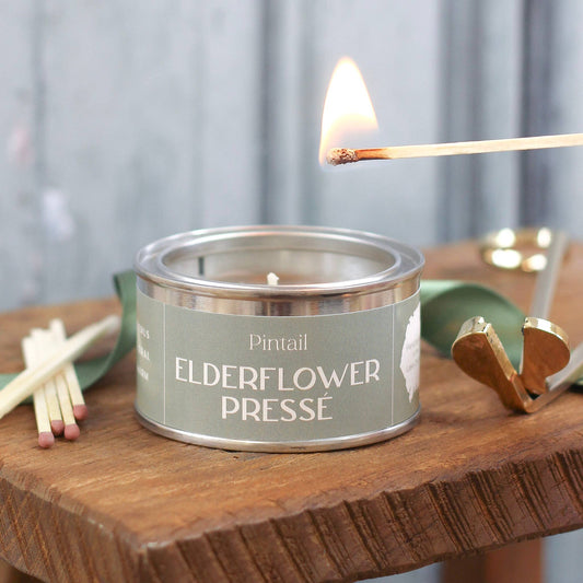 Candle labeled 'Pintail Elderflower Presse' being lit with a match on a wooden surface.
