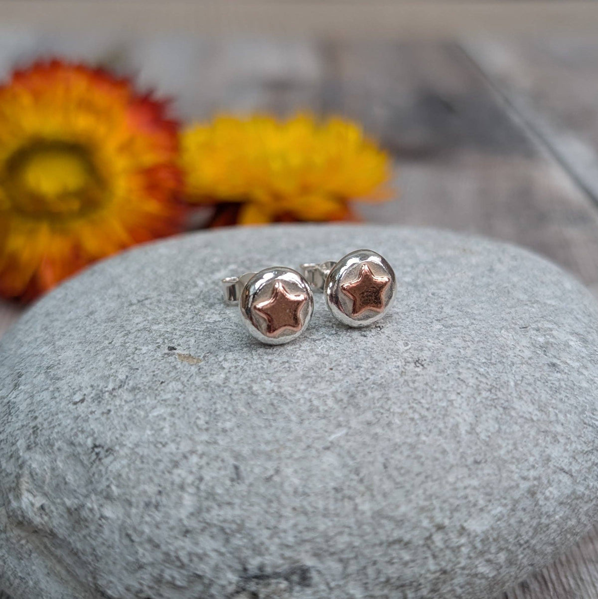 A pair of silver stud earrings with star-shaped cutouts, displayed on a smooth gray stone with blurred yellow and orange flowers in the background.