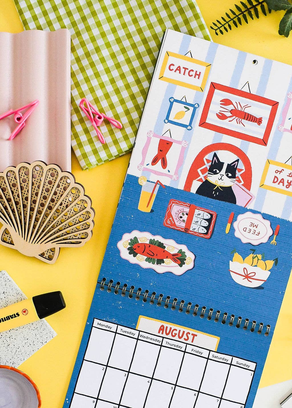 A spiral-bound monthly calendar page with a blue background and colorful fishing-themed illustrations, photographed on a bright yellow surface with other styled props including a green gingham cloth, pink clothespins, a wooden seashell object, and a small notebook.