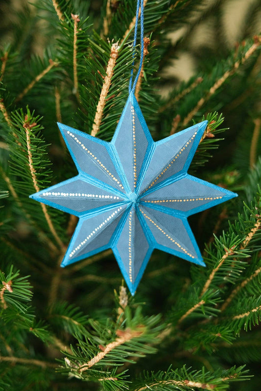 A light blue star-shaped Christmas ornament with gold glitter accents hanging from a blue string in the green needles of a Christmas tree.