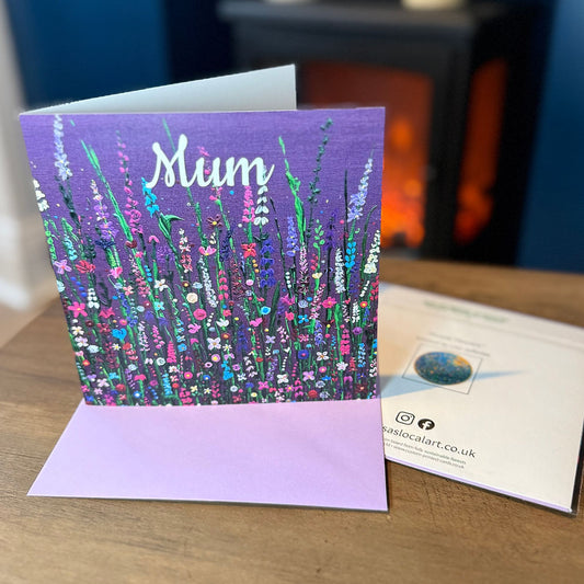 A square greeting card with a purple background and painted colourful wildflowers, with the word 'Mum' in white script on the top. The card is shown standing open on a wooden table next to its lavender envelope.