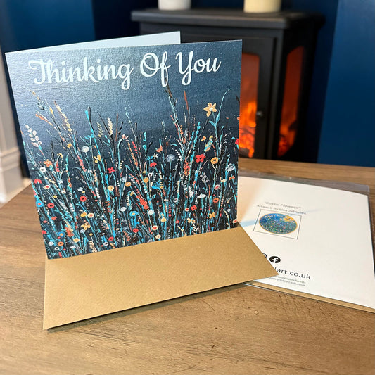 A greeting card standing on a wooden table, printed with the words 'Thinking Of You' above a colorful wildflower illustration against a dark background.