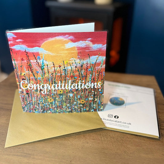 A square card featuring a colourful painted field of flowers under a yellow sun and red sky, with the word 'Congratulations' written across the front. It is displayed on a wooden table next to a gold envelope and a printed card.