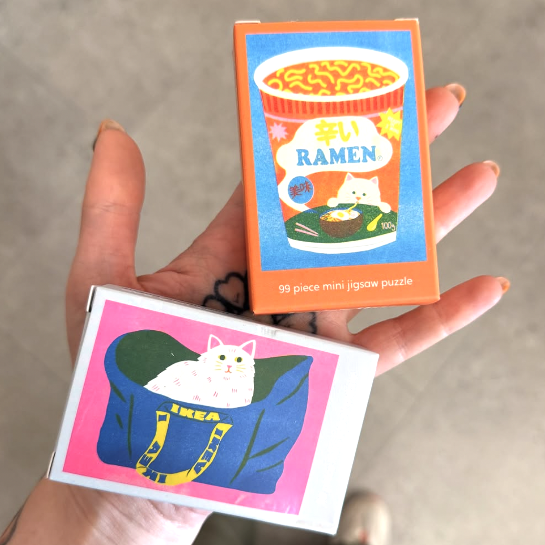 Two mini jigsaw puzzle boxes held in a hand, one with a cat in a bag design and the other with a ramen cup design.