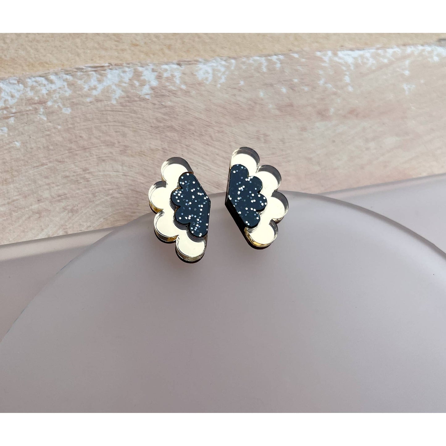 A pair of small cloud-shaped stud earrings with a dark navy glitter center, cream enamel edges, and gold-tone outlines, displayed on a round gray stand.