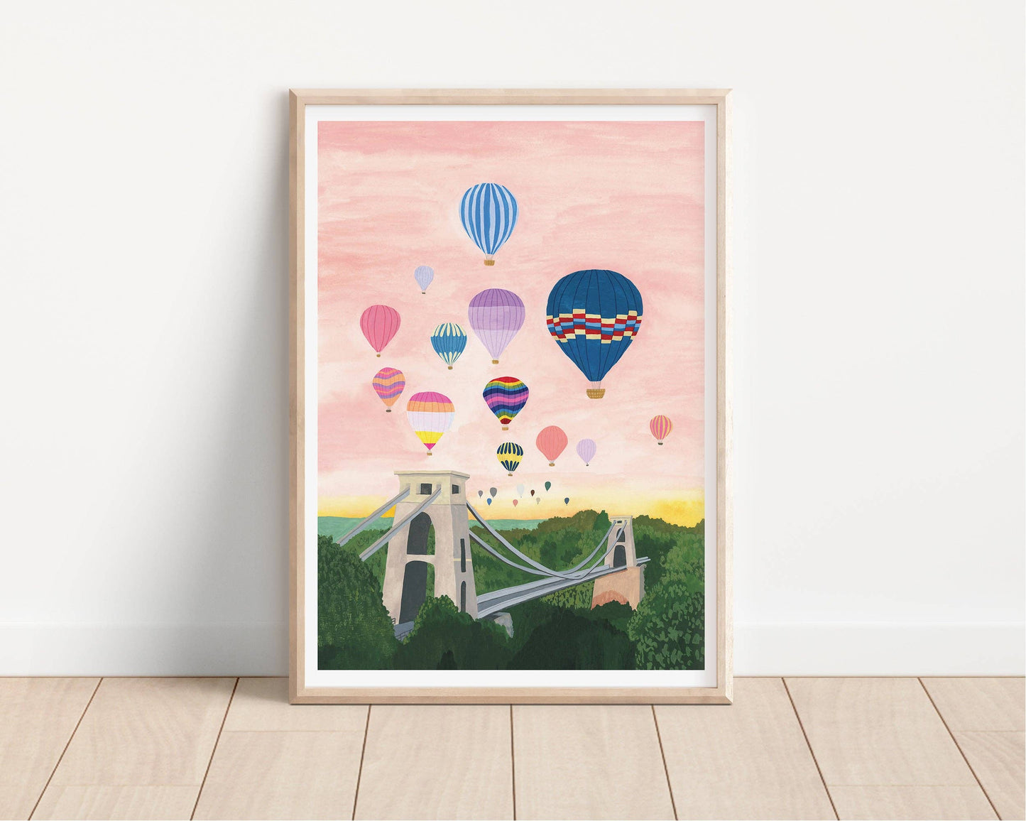 Framed artwork of colorful hot air balloons over a bridge on a pink sky background.