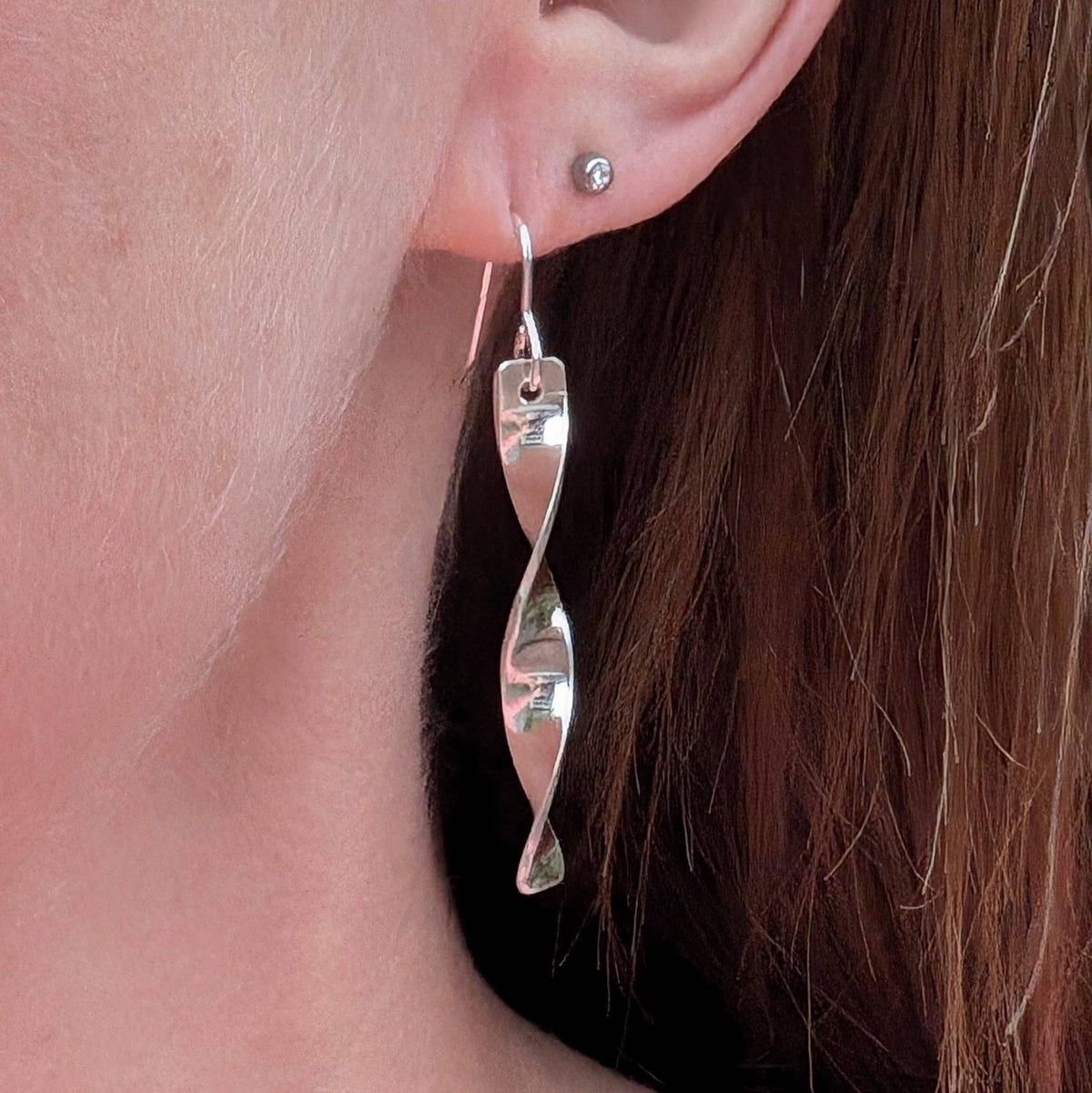 A close-up of a person's ear wearing a long, twisted silver dangle earring, with a small round stud earring on the opposite ear and brown hair visible.