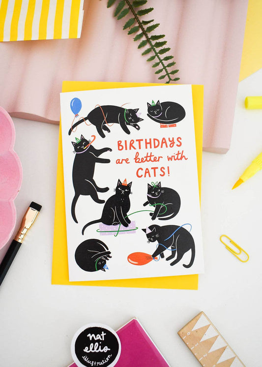 Greeting card with black cats and text 'Birthdays are better with cats!' on a white surface with stationery items.