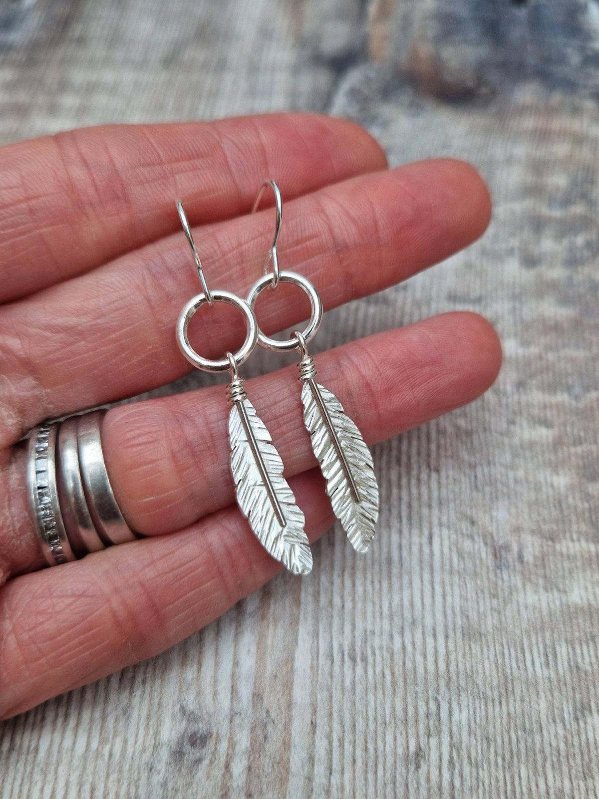 A pair of silver feather drop earrings hanging from a person's fingers, with a silver ring on one finger, set against a rustic wooden background.