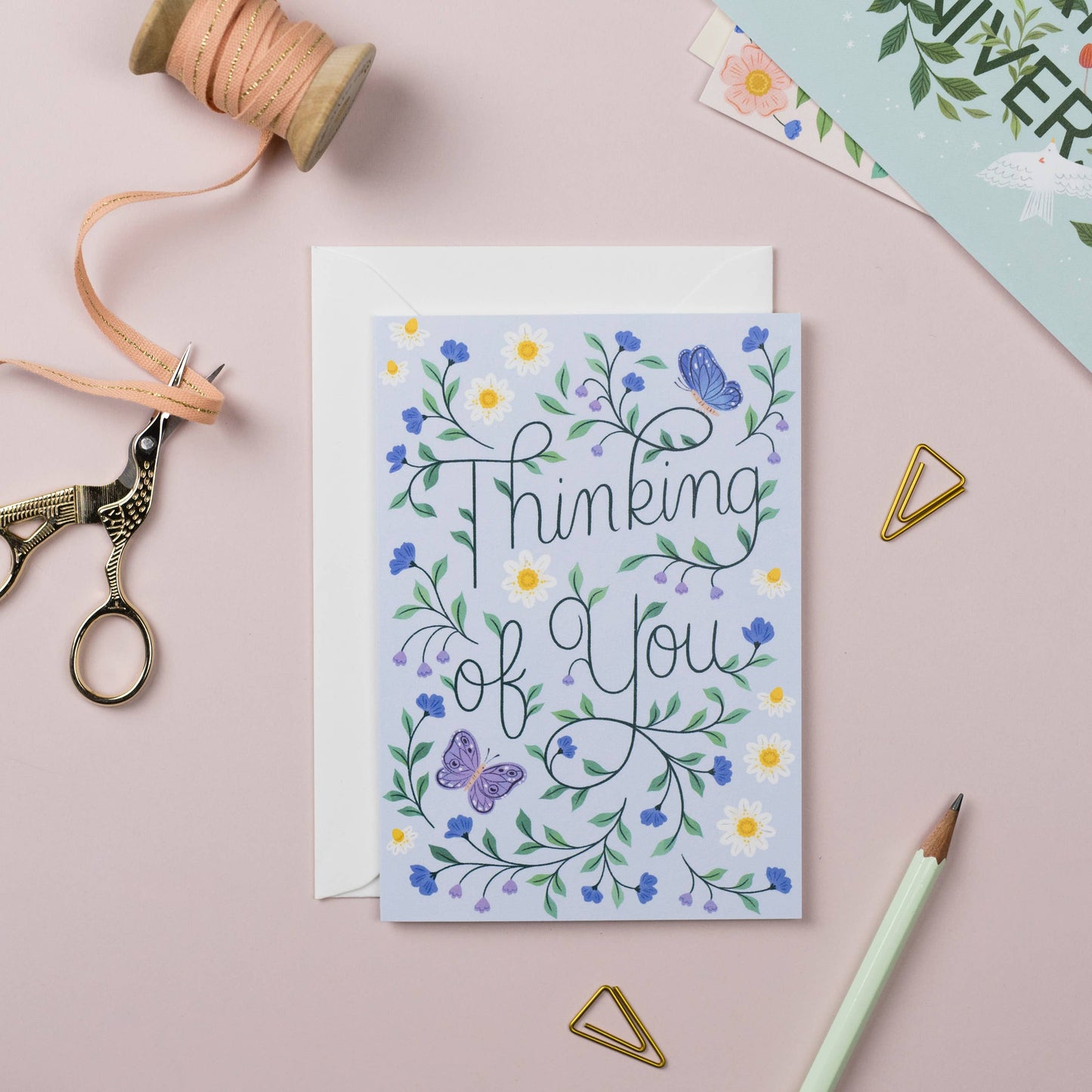 Greeting card with floral design and 'Thinking of You' text on a pink surface with crafting supplies.