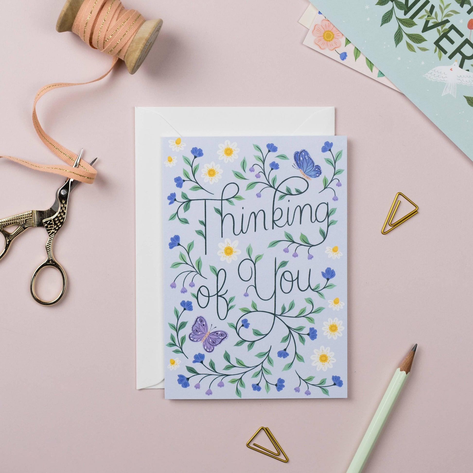 Greeting card with floral design and 'Thinking of You' text on a pink surface with crafting supplies.