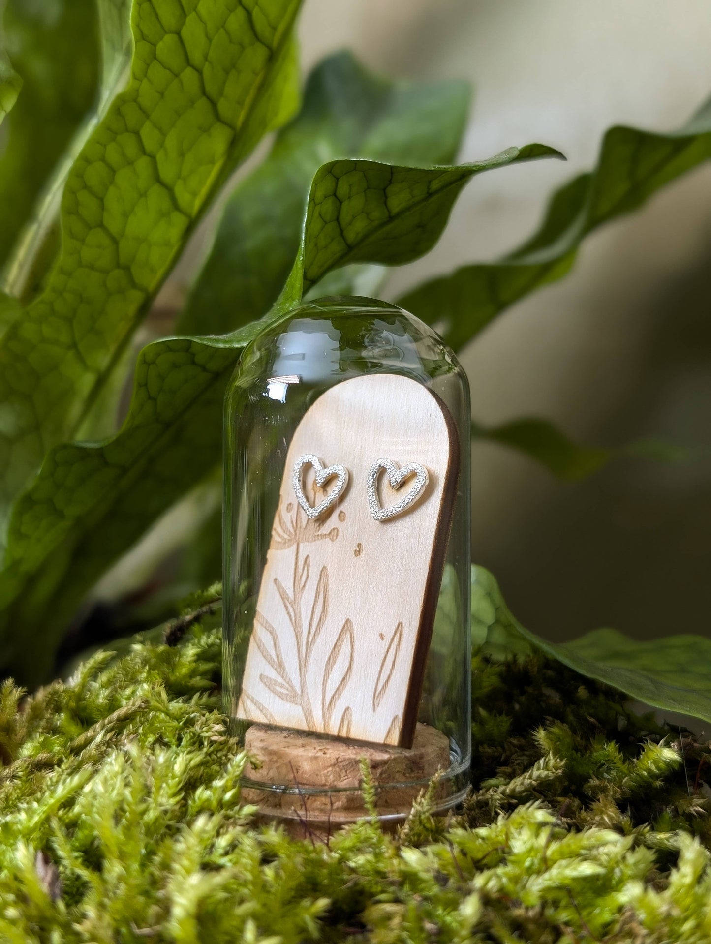 Sterling silver heart-shaped stud earrings with a textured design, presented in a glass terrarium tube, placed on a mossy surface with green leaves in the background.