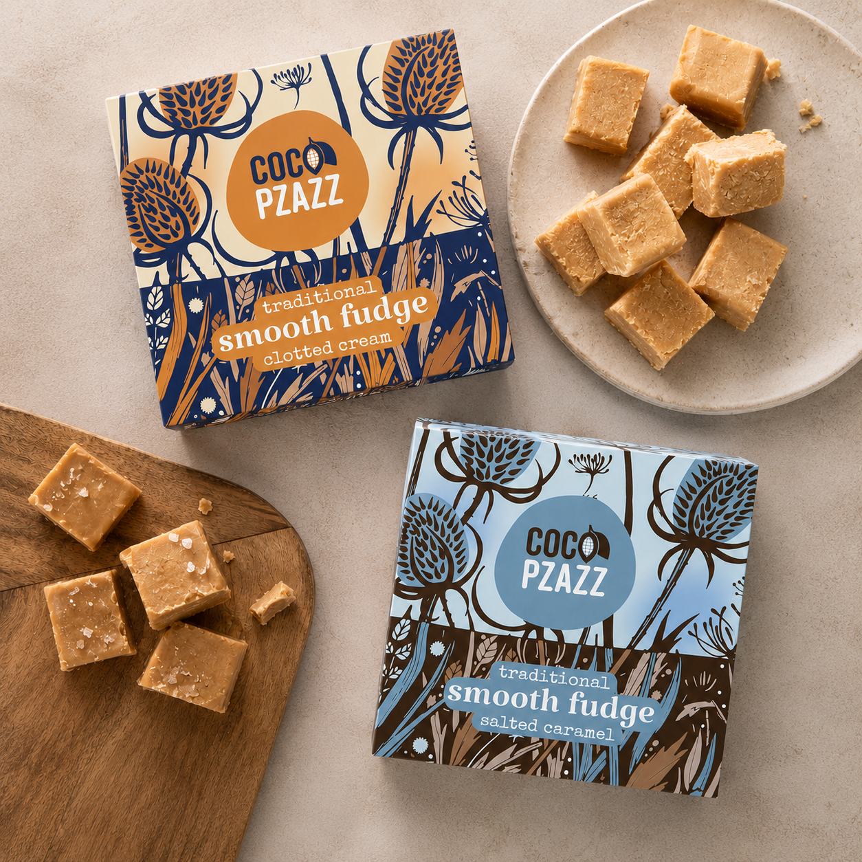 Two boxes of Coco Pzaazz chocolate fudge arranged on a beige surface, with some fudge cubes displayed on a wooden board and a plate. The boxes feature botanical illustrations in navy and tan, with one box showing a blue circle logo and the other showing an orange circle logo.