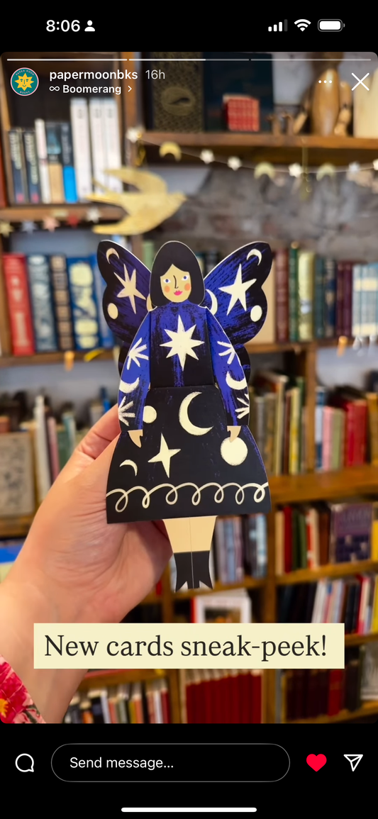 Hand holding a paper fairy card with star and moon designs in front of a bookshelf.