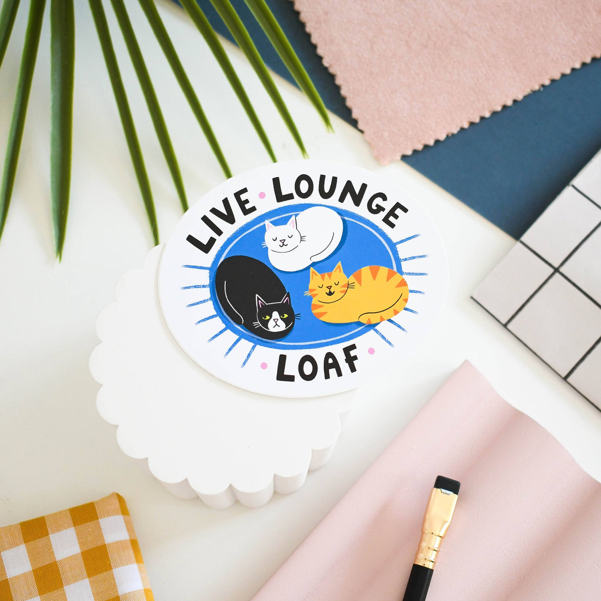 A round sticker with a scalloped white border featuring the text 'LIVE LOUNGE LOAF' and illustrations of three cats (black, white, and orange) on a blue background, displayed on a styled flat-lay surface with a plant leaf, pink fabric, and a paintbrush.