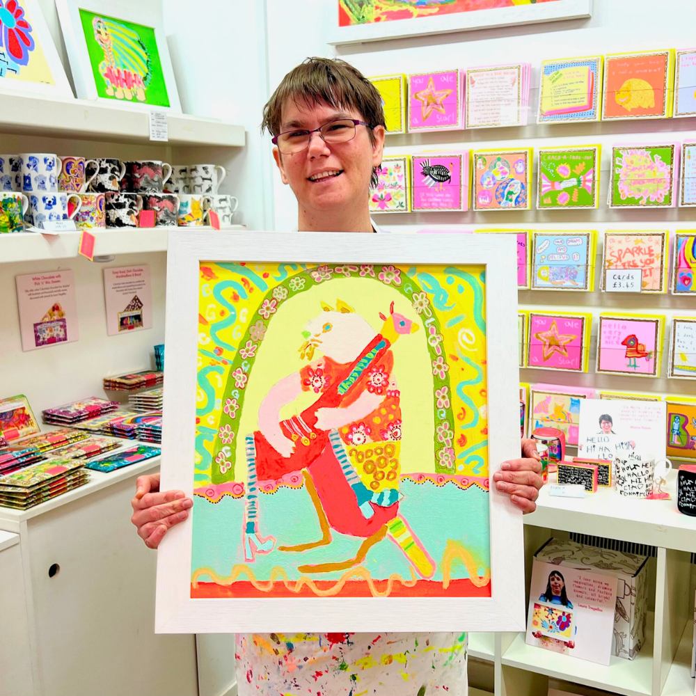 Person holding a colorful framed artwork in a store with various items on shelves.