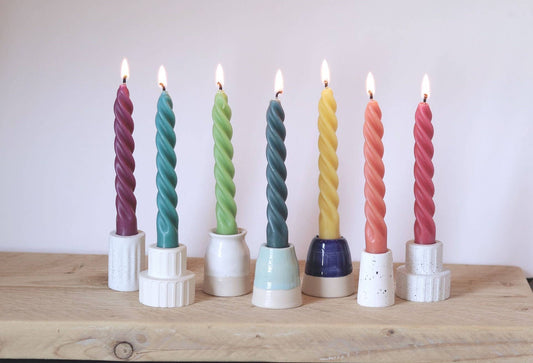 Seven twisted taper candles in assorted colors standing in white ceramic holders on a light wood surface against a plain light background.