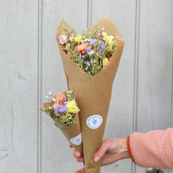 Bouquet of flowers wrapped in brown paper with a visible brand logo, held against a light wooden background.