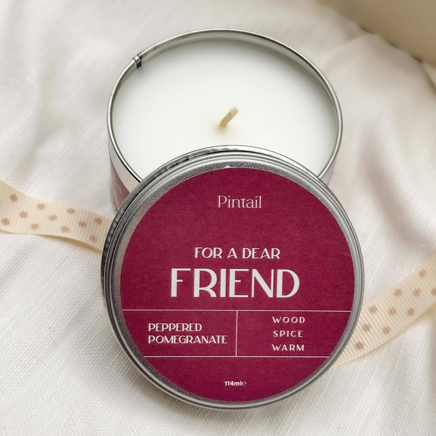 Candle in a tin labeled 'For a Dear Friend' by Pintail on a white fabric background.