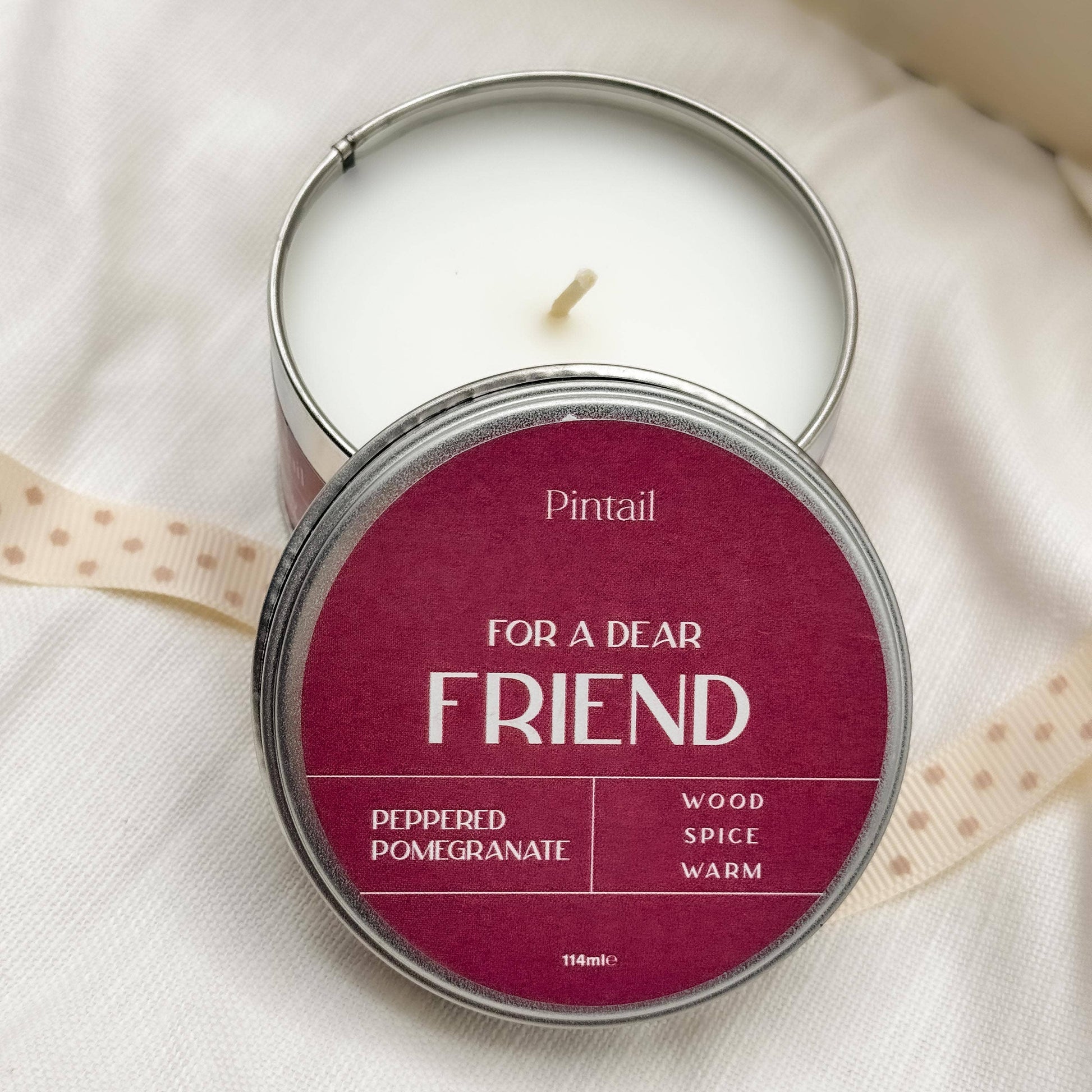 Candle in a tin labeled 'For a Dear Friend' by Pintail on a white fabric background.