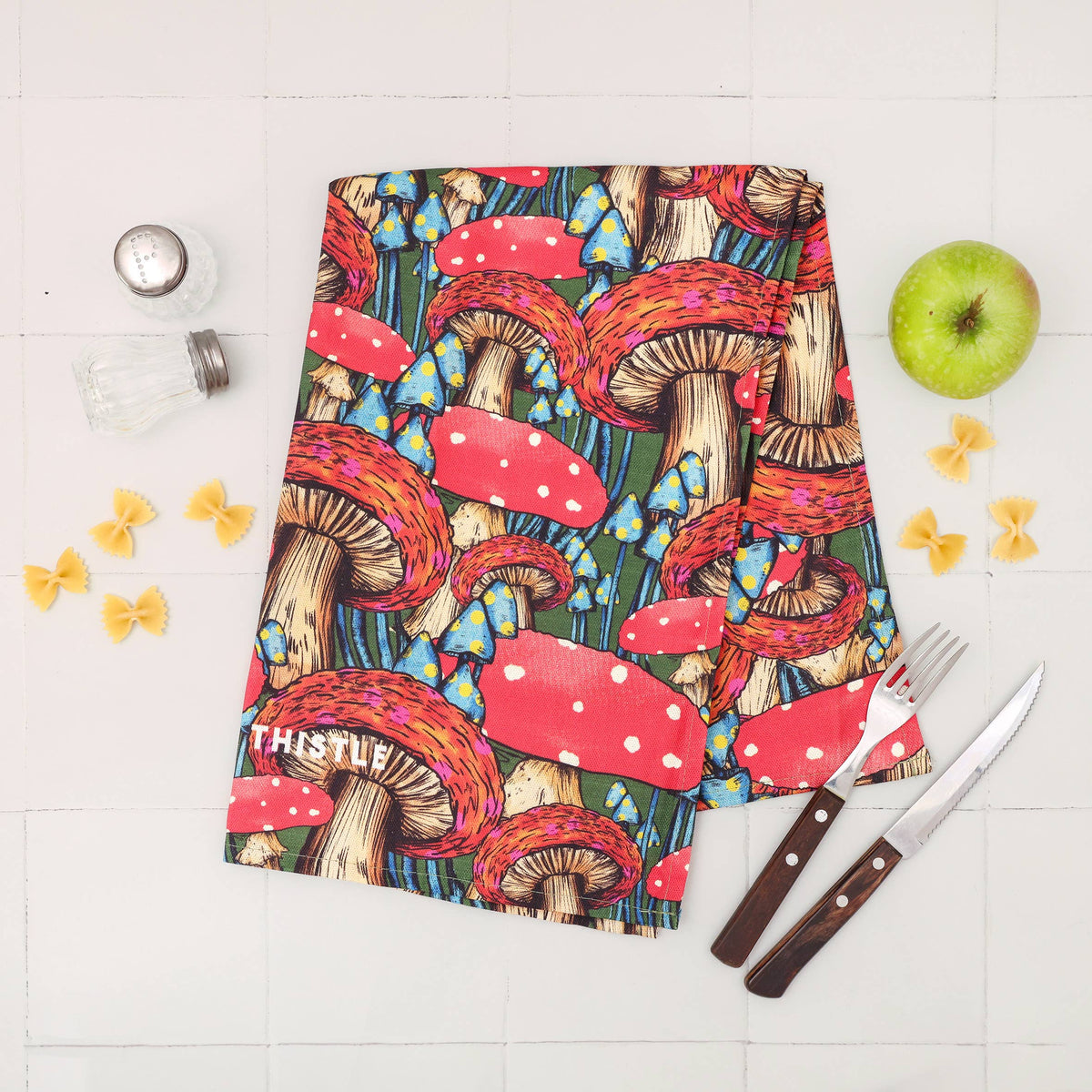 A square kitchen towel with a colorful mushroom print is laid flat on a white tiled surface, styled with a green apple, pasta, a salt shaker, and a fork and knife.