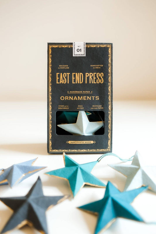 A boxed set of paper star ornaments by East End Press, with the black-and-gold package standing upright and several star ornaments in teal, gray, and white arranged in front on a light beige surface.
