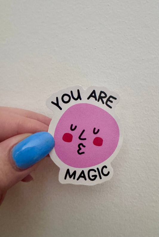 Sticker with a pink circle and text held by a hand with blue nail polish on a light gray background
