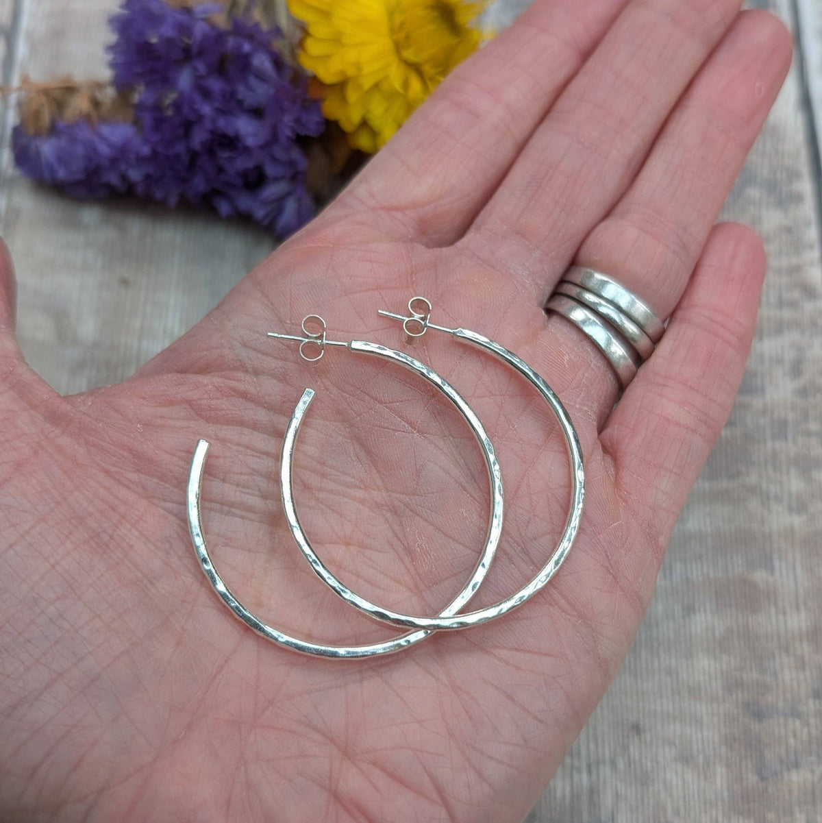 A pair of hammered silver hoop earrings displayed on an open hand, with a silver ring on the model's finger and dried purple and yellow flowers in the background.