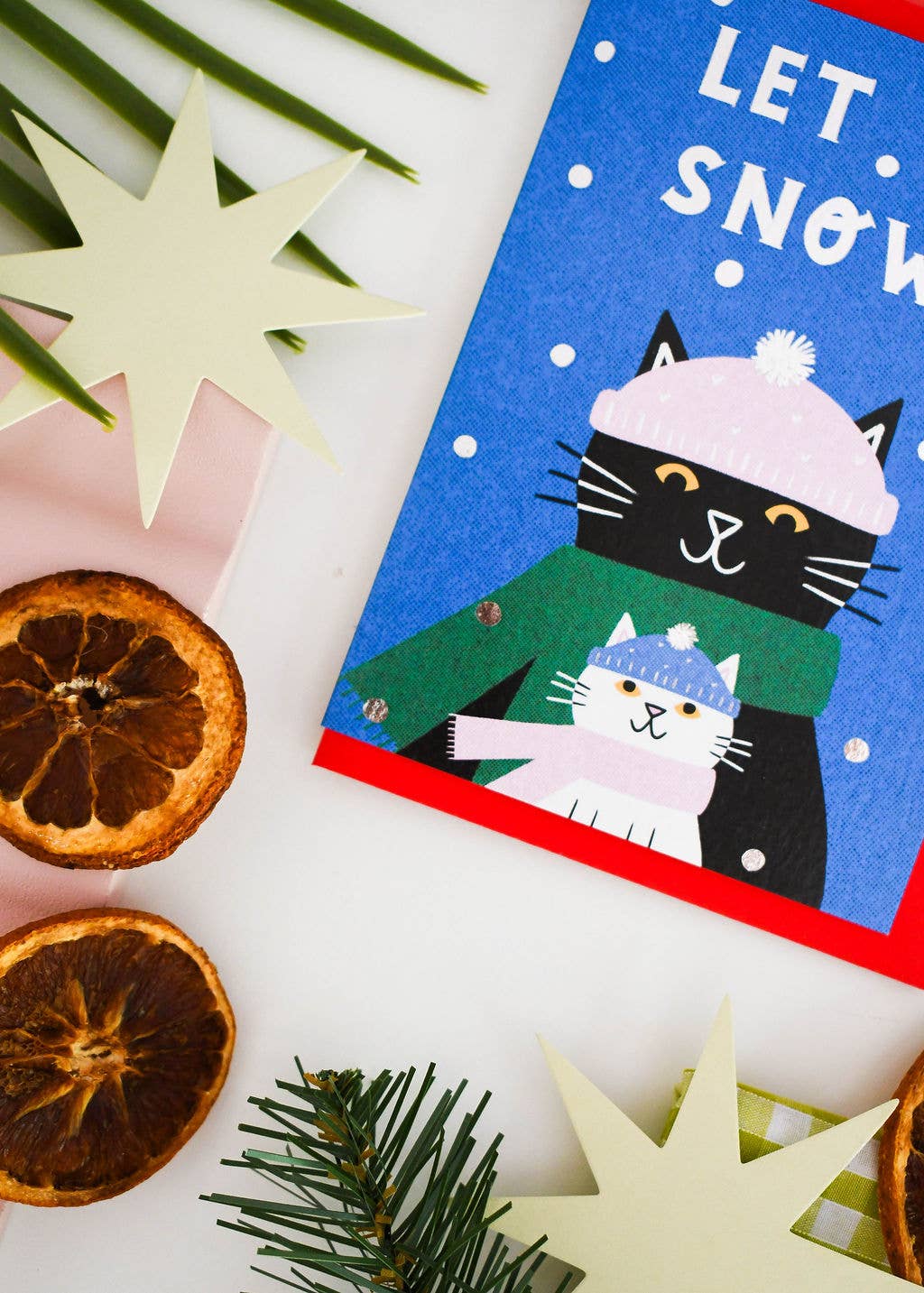 A festive holiday greeting card featuring a black cat in a pink hat and green scarf hugging a white cat, with the text 'LET SNOW W.