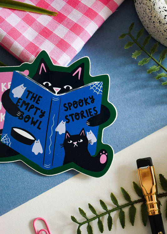 A die-cut sticker featuring a black cat reading a blue book titled 'The Empty Bowl: Spooky Stories' is displayed on a styled flat-lay background with a pink gingham cloth, blue paper, green leaves, and a gold pen.