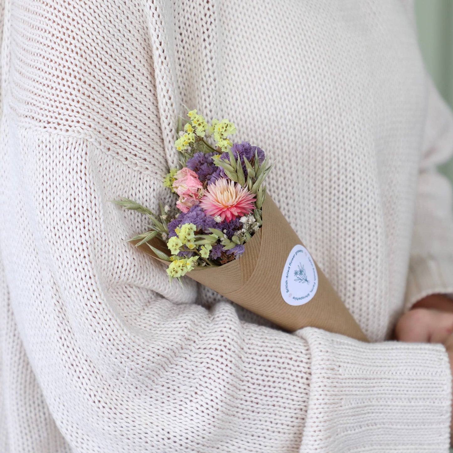 Person holding a small bouquet of flowers wrapped in brown paper with a white label against a neutral background.