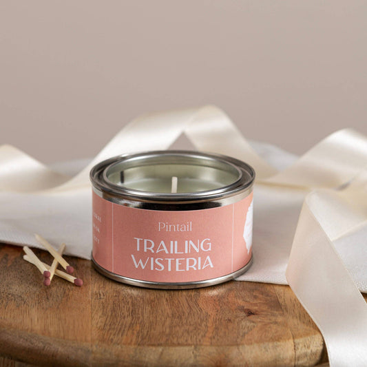 Candle in a tin labeled 'Trailing Wisteria' by Pintail on a wooden surface with white fabric.