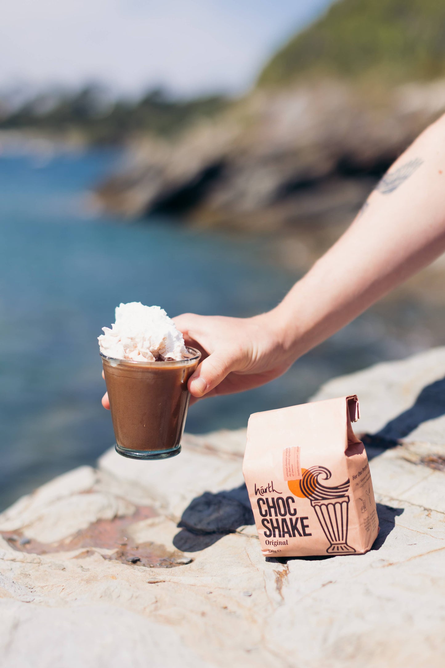 Hand holding a Choc Shake with whipped cream by a scenic coastal background