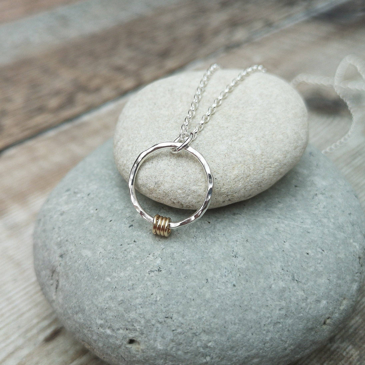 A silver ring pendant necklace with a small gold wire accent, resting on smooth gray stones over a rustic wooden surface.