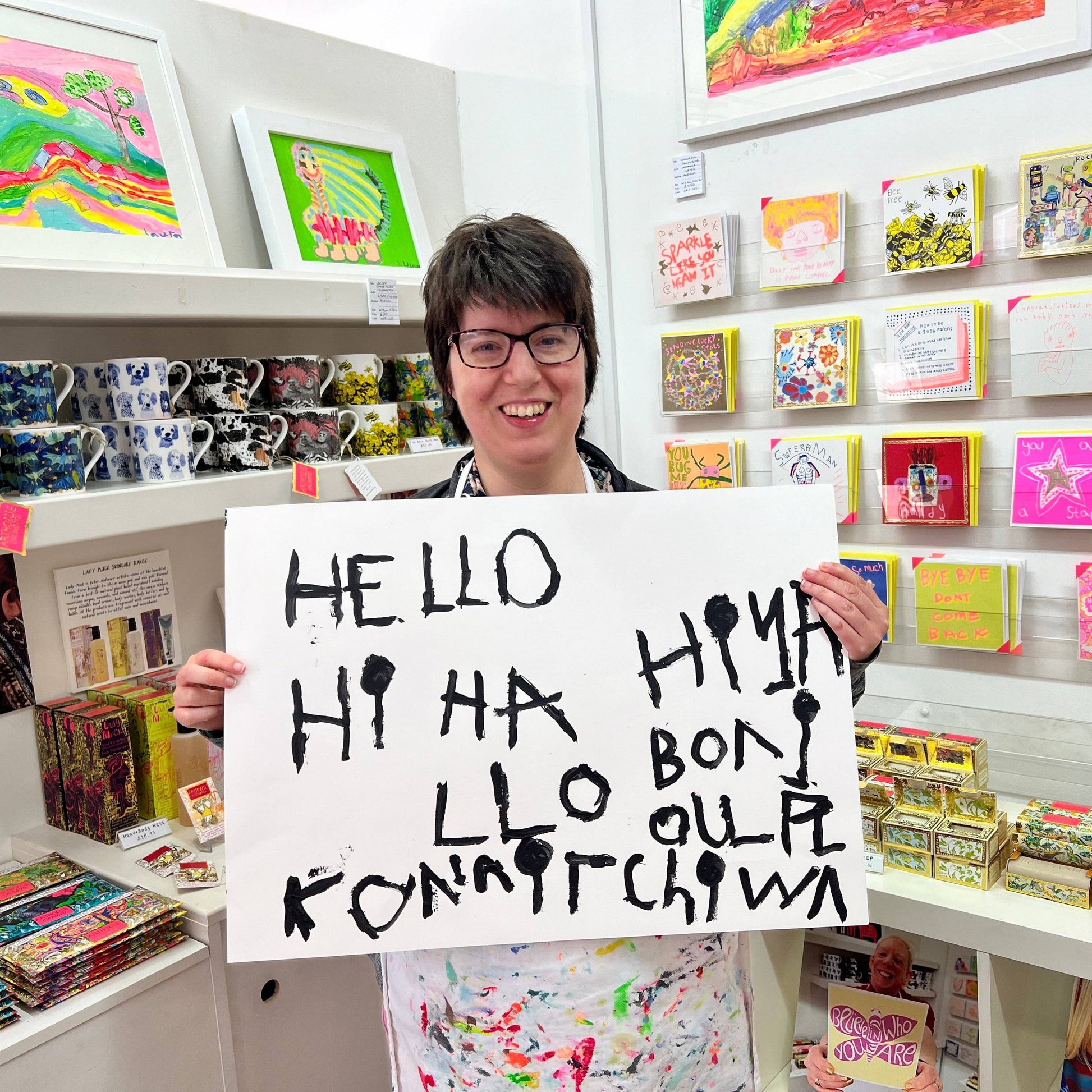 Person holding a sign with 'Hello' in multiple languages in a store with shelves of products.