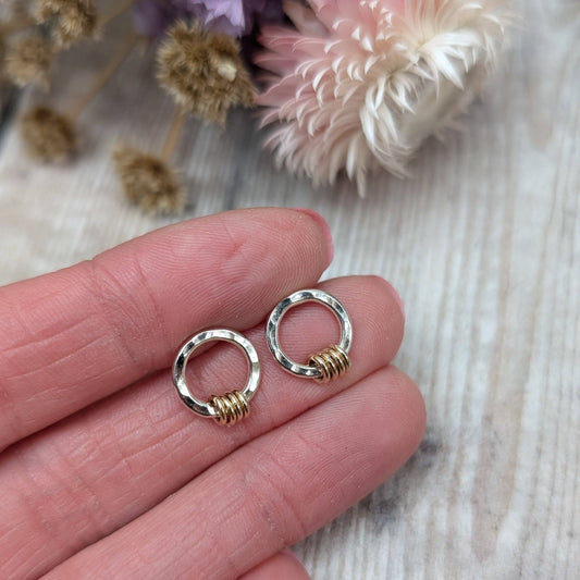 A pair of small hoop earrings with textured silver tops and gold spiral wraps, held on a person's fingers against a light wood background with dried flowers.