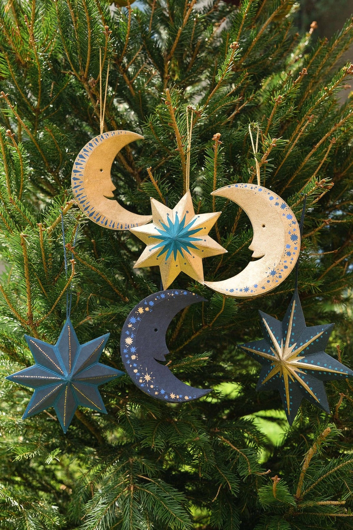 Hand-painted wooden Christmas ornaments shaped like crescent moons and stars hanging on a green pine tree. The ornaments feature natural wood tones with blue and yellow painted details.