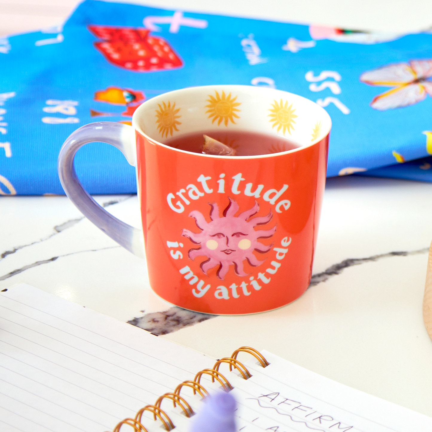 Red mug with 'Gratitude is my attitude' text on a marble surface with a colorful fabric background.
