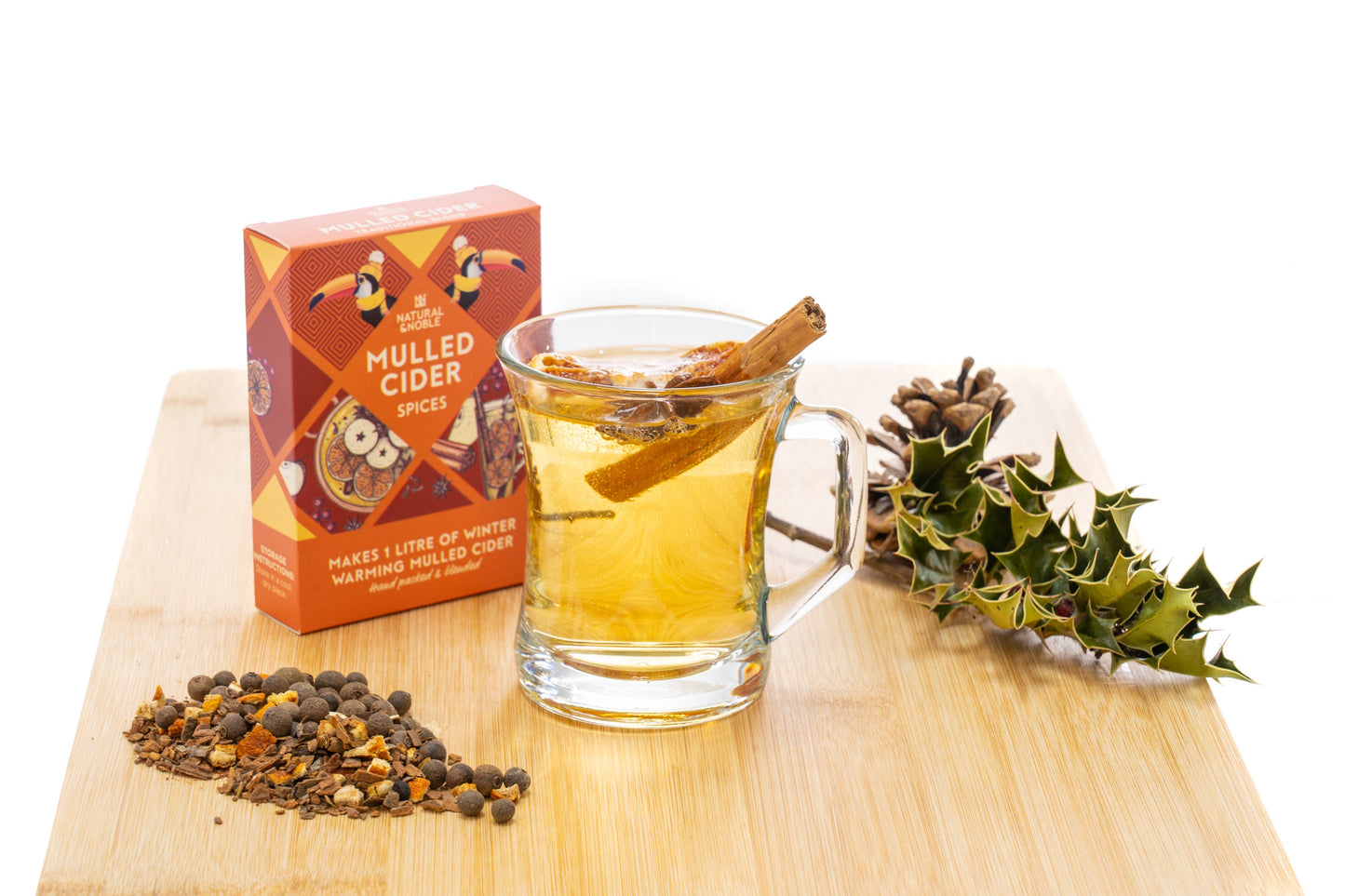 Glass of mulled cider with cinnamon sticks on a wooden board, next to a box of mulled cider spices and holly leaves.