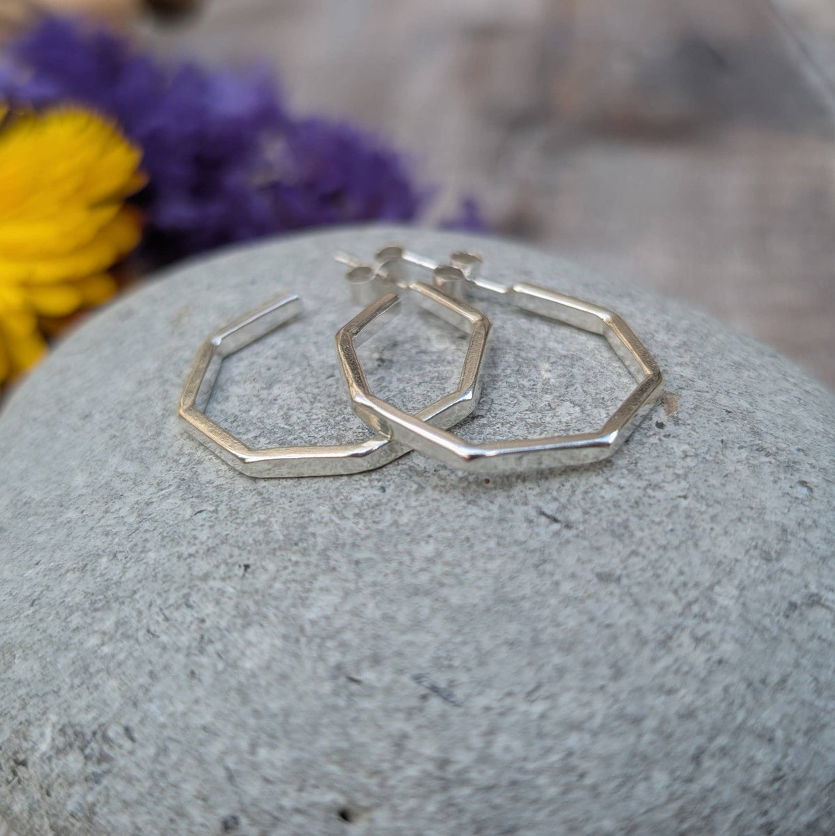 A pair of silver-toned geometric hoop earrings with a hexagonal open design, resting on a smooth gray stone with blurred purple and yellow flowers in the background.