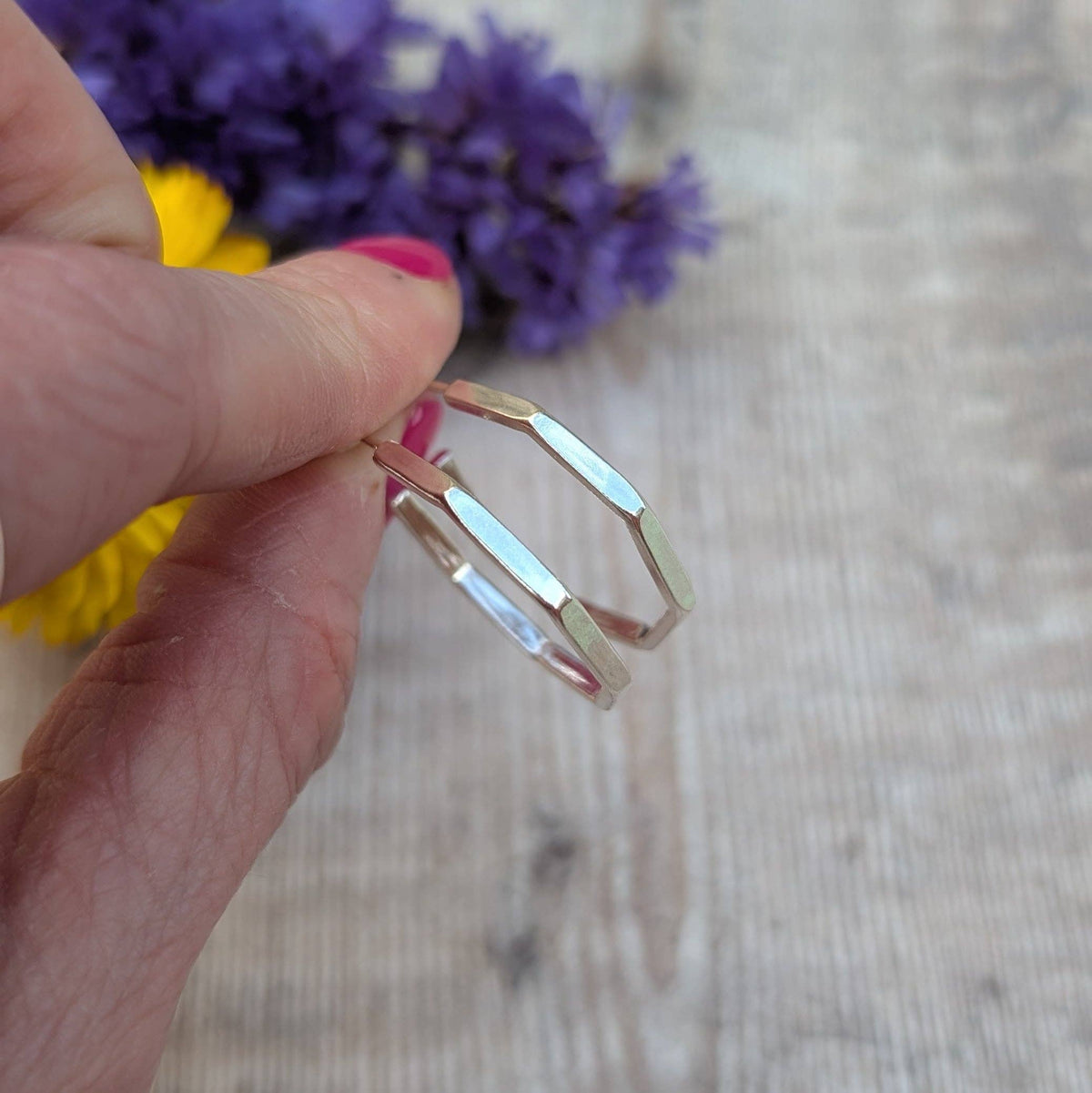 A hand with pink nail polish holds a silver-toned geometric bangle bracelet with elongated rectangular links, set against a light wood background with blurred purple and yellow flowers.