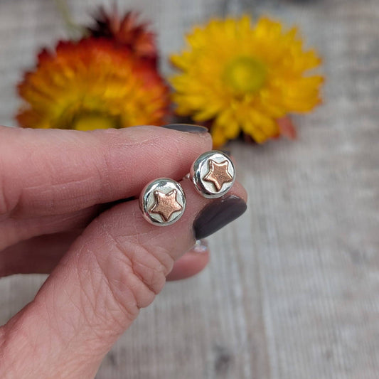 A close-up of a hand holding a pair of small round stud earrings with star-shaped details, one in silver and one in rose gold, against a blurred background with yellow and orange flowers.